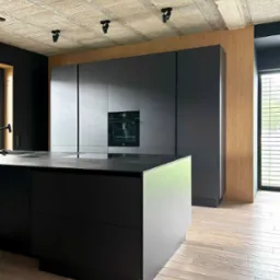 Nowoczesna kuchnia: czarne, matowe fronty szafek, wyspa kuchenna z blatem z ciemnego kamienia, surowy betonowy sufit z reflektorami, drewniana okładzina ściany i jasna drewniana podłoga.