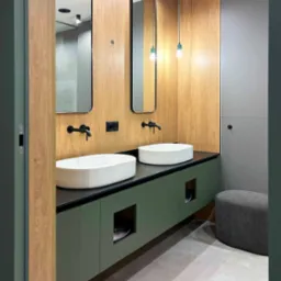 Nowoczesna łazienka z dwiema owalnymi umywalkami nablatowymi, czarną armaturą, szafką w odcieniach zieleni, drewnianą boazerią na ścianie i wiszącymi lampami z żarówkami edisona.