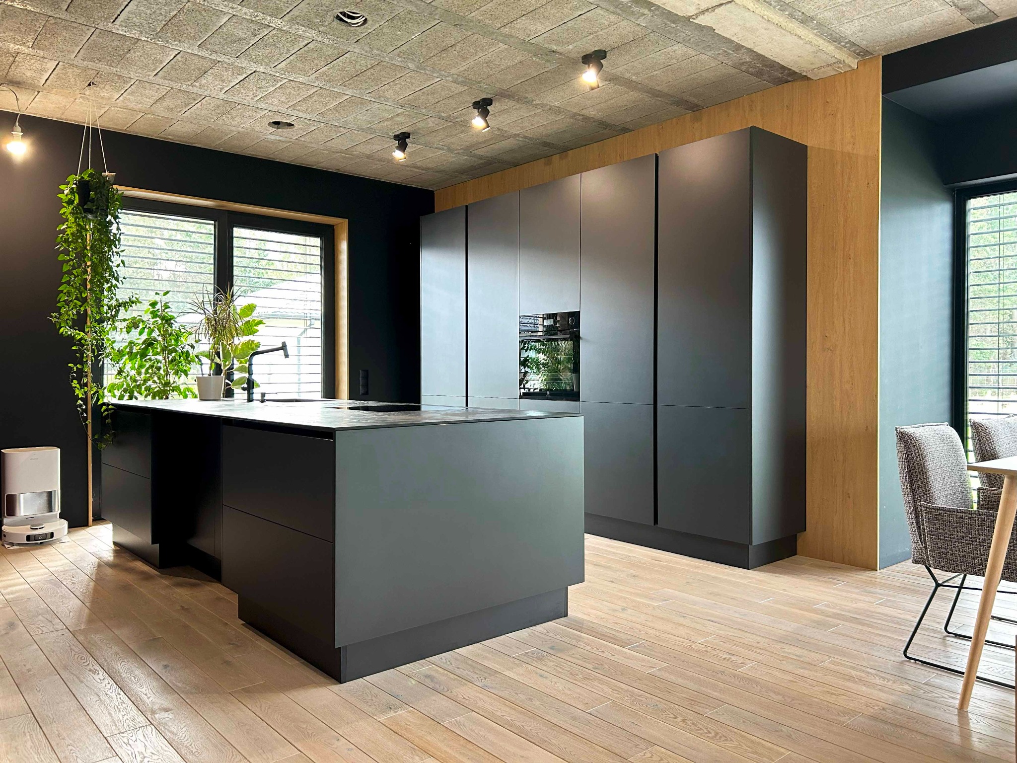 Nowoczesna, minimalistyczna kuchnia w odcieniach szarości z wyspą, betonowym sufitem i drewnianą podłogą. Na wyspie widoczne rośliny doniczkowe, a obok okna zwisająca zieleń. W tle zabudowa...