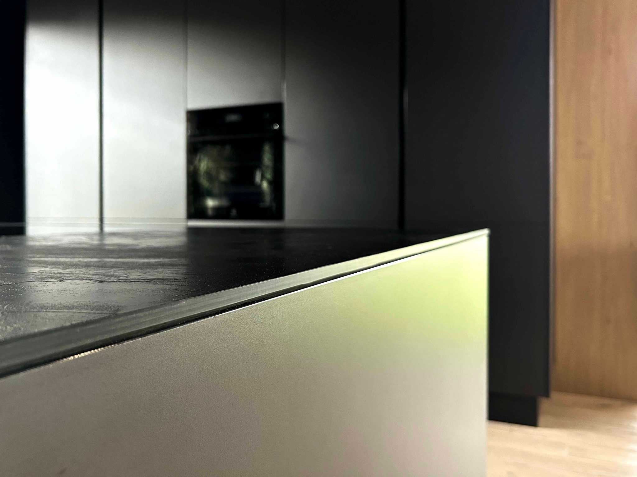 Szczegółowe ujęcie krawędzi blatu kuchennego z ciemnego kamienia, kontrastującego z jasnym frontem szafki; w tle zabudowa kuchenna z piekarnikiem.