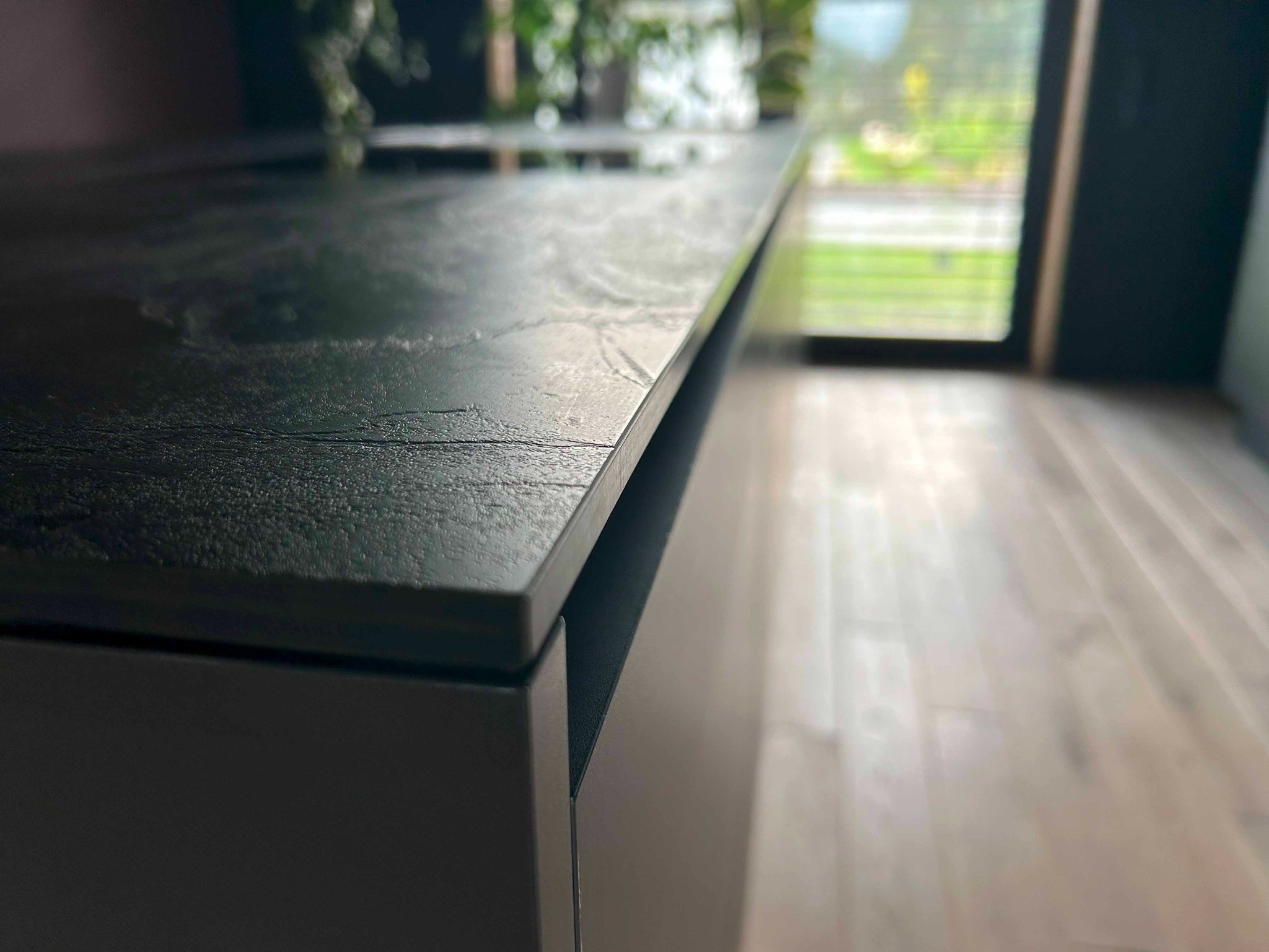 Czarny blat kuchenny z wyraźną teksturą, widoczny z bliska, z oknem w tle i roślinami na górze, oświetlony naturalnym światłem.