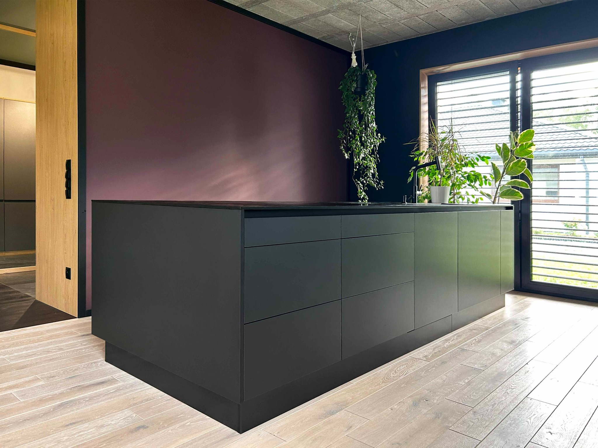 Nowoczesna, matowa, grafitowa wyspa kuchenna z blatem w kolorze czarnym, na jasnej, drewnianej podłodze, z wiszącą rośliną doniczkową i widokiem na ogród przez okno z żaluzjami.