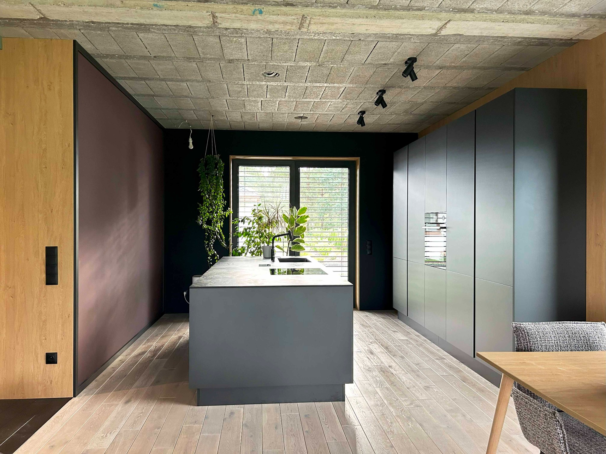 Nowoczesna kuchnia: szare szafki, wyspa z blatem, betonowy sufit z oświetleniem punktowym, drewniane podłogi i rośliny doniczkowe przy oknie.
