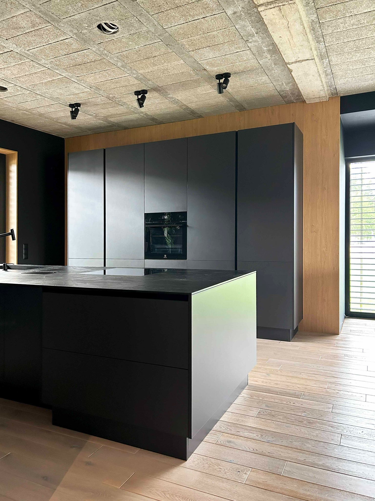 Nowoczesna kuchnia: czarne, matowe fronty szafek, wyspa kuchenna z blatem z ciemnego kamienia, surowy betonowy sufit z reflektorami, drewniana okładzina ściany i jasna drewniana podłoga.
