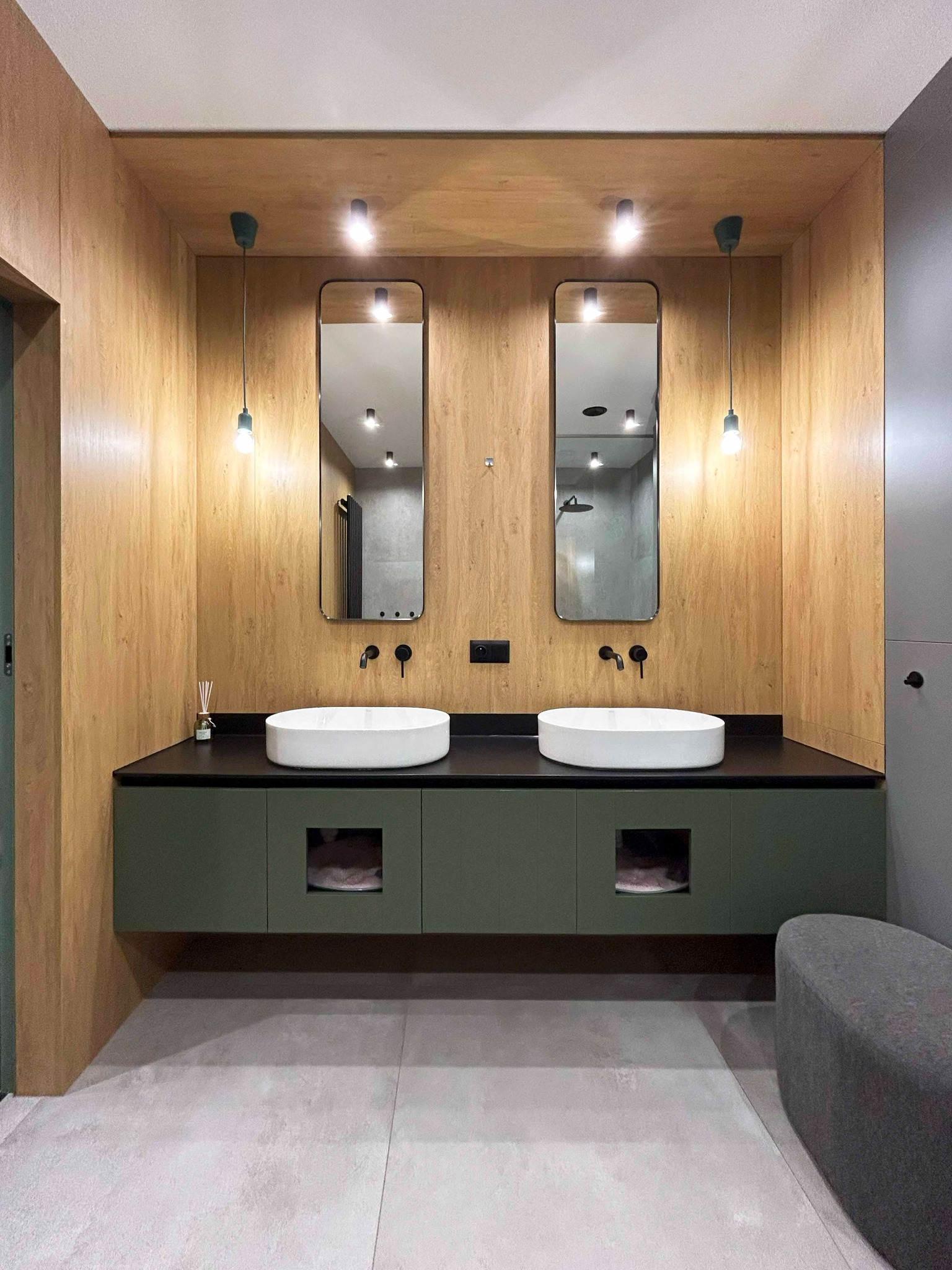 Nowoczesna łazienka z dwoma białymi umywalkami nablatowymi, czarnym blatem i szafką w odcieniach zieleni, drewniane panele ścienne i dwa lustra z czarnymi ramami oraz wiszące lampy z żarówkami.