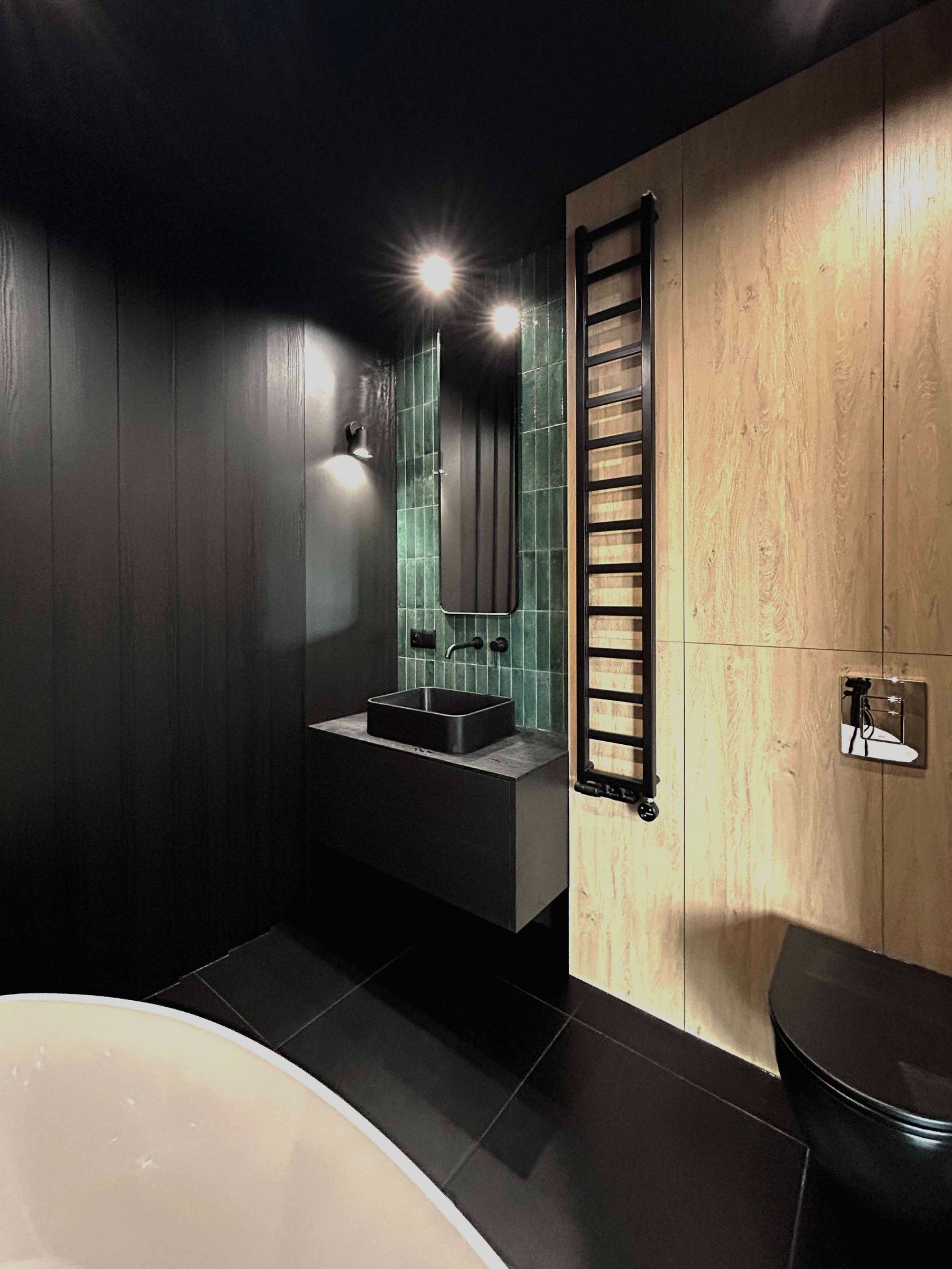 Nowoczesna łazienka z czarnymi ścianami, drewnianą zabudową i zielonymi płytkami, z czarną umywalką nablatową i grzejnikiem drabinkowym.