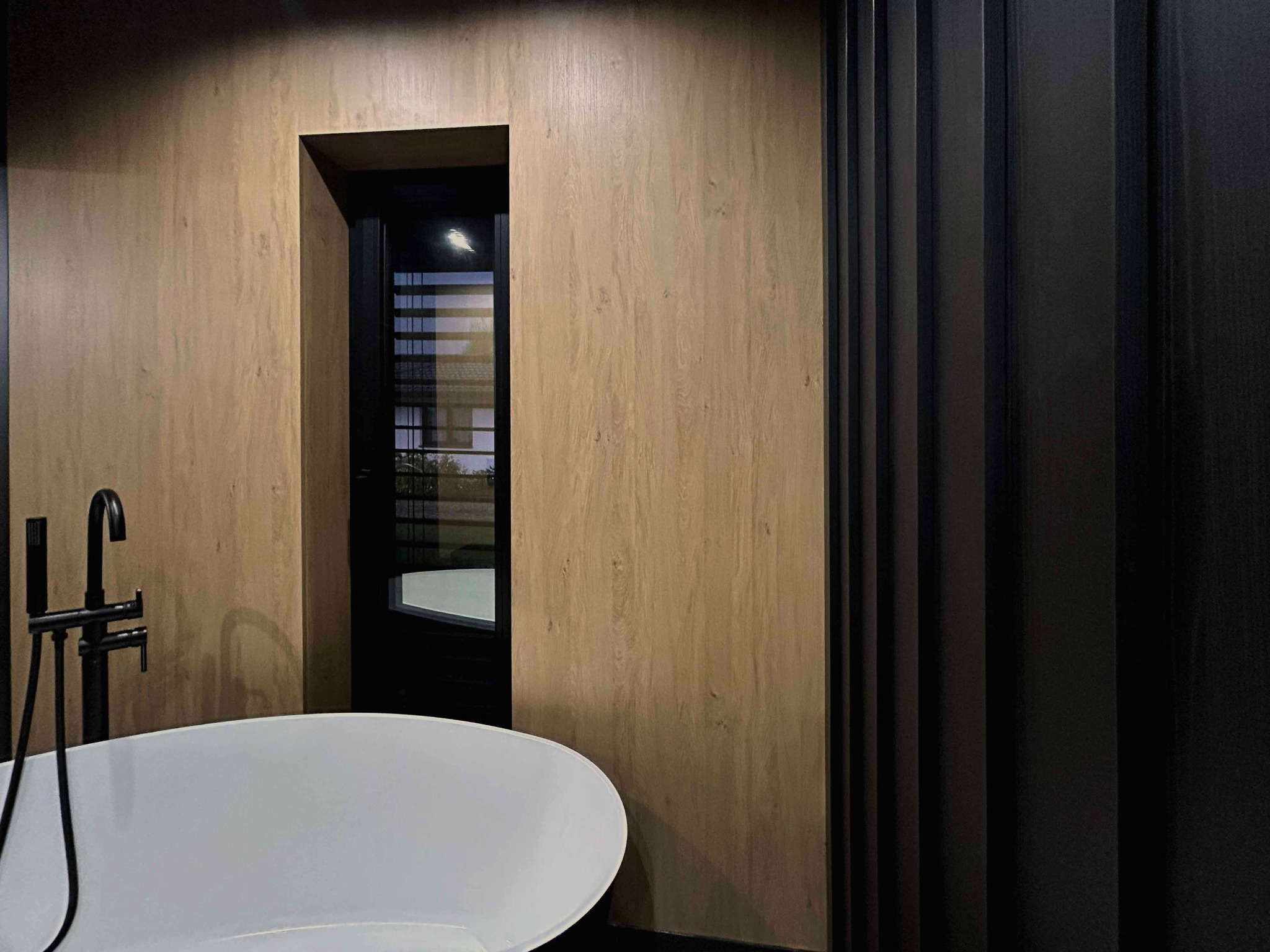 Fragment łazienki z wolnostojącą białą wanną, czarną armaturą i ścianą wykończoną panelami imitującymi drewno, widoczne czarne pionowe lamele dekoracyjne.