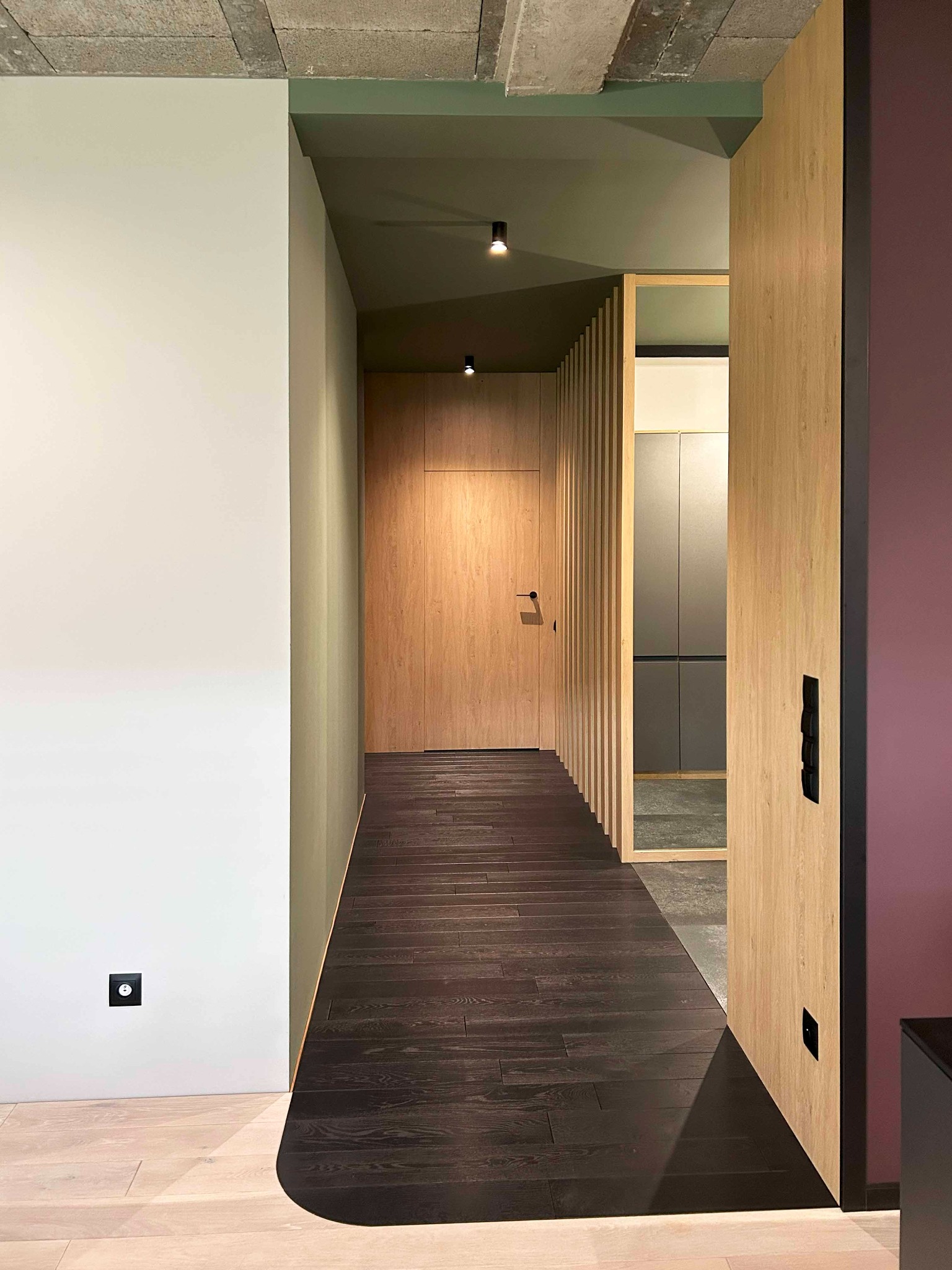 Nowoczesny korytarz z ciemną drewnianą podłogą, jasnymi drewnianymi drzwiami przesuwnymi z czarnymi uchwytami, ścianą w kolorze złamanej bieli i surowym betonowym sufitem z punktowym oświetleniem.