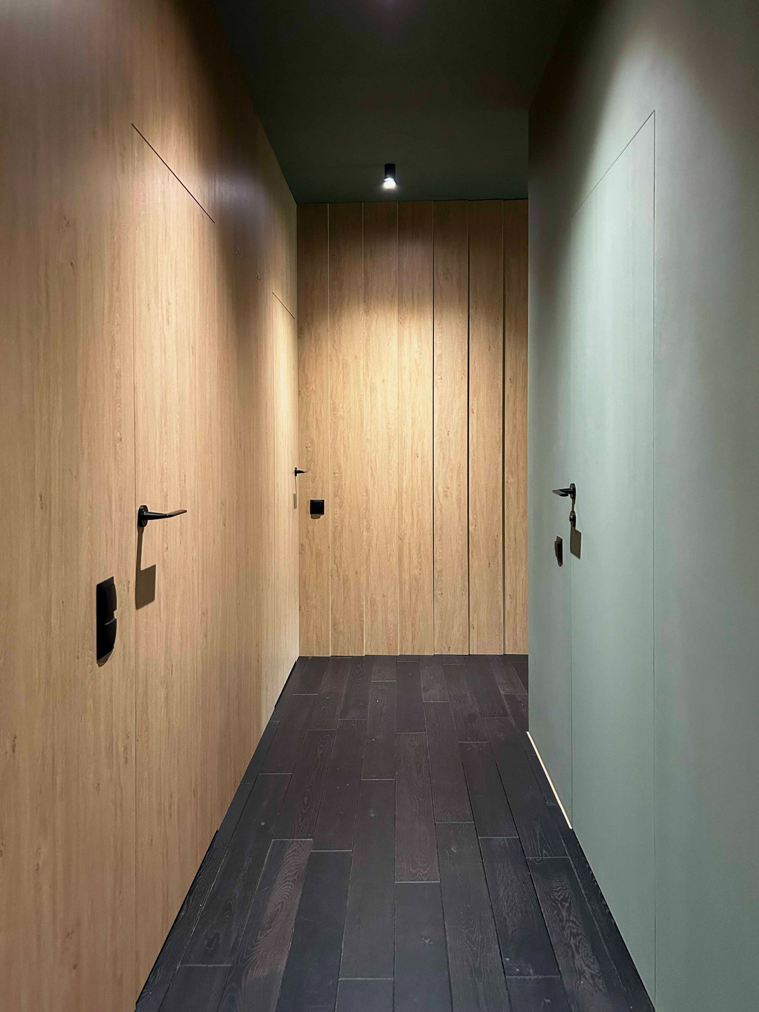 Wąski korytarz z zabudową szaf wnękowych z ukrytymi drzwiami, wykończonych drewnopodobną okleiną, czarne klamki i ciemna, drewniana podłoga.