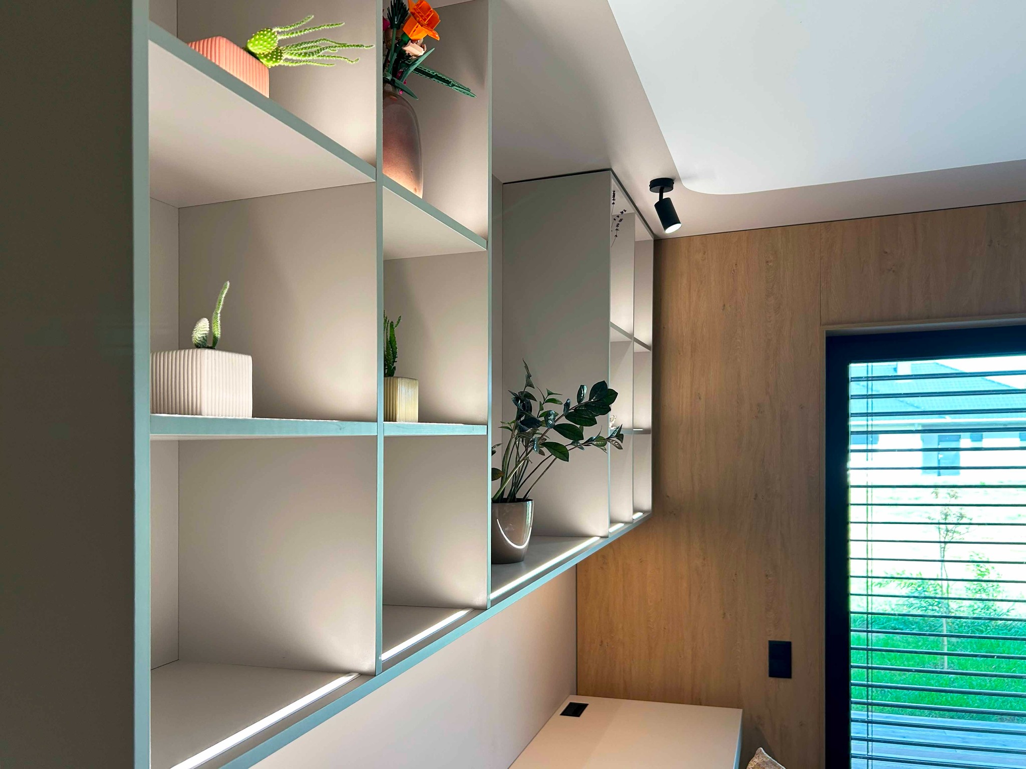 Nowoczesna, jasna zabudowa meblowa z półkami w Łańcucie, z dekoracyjnymi roślinami w doniczkach, oświetlona punktowymi lampami, na tle drewnianej ściany i okna z żaluzjami.