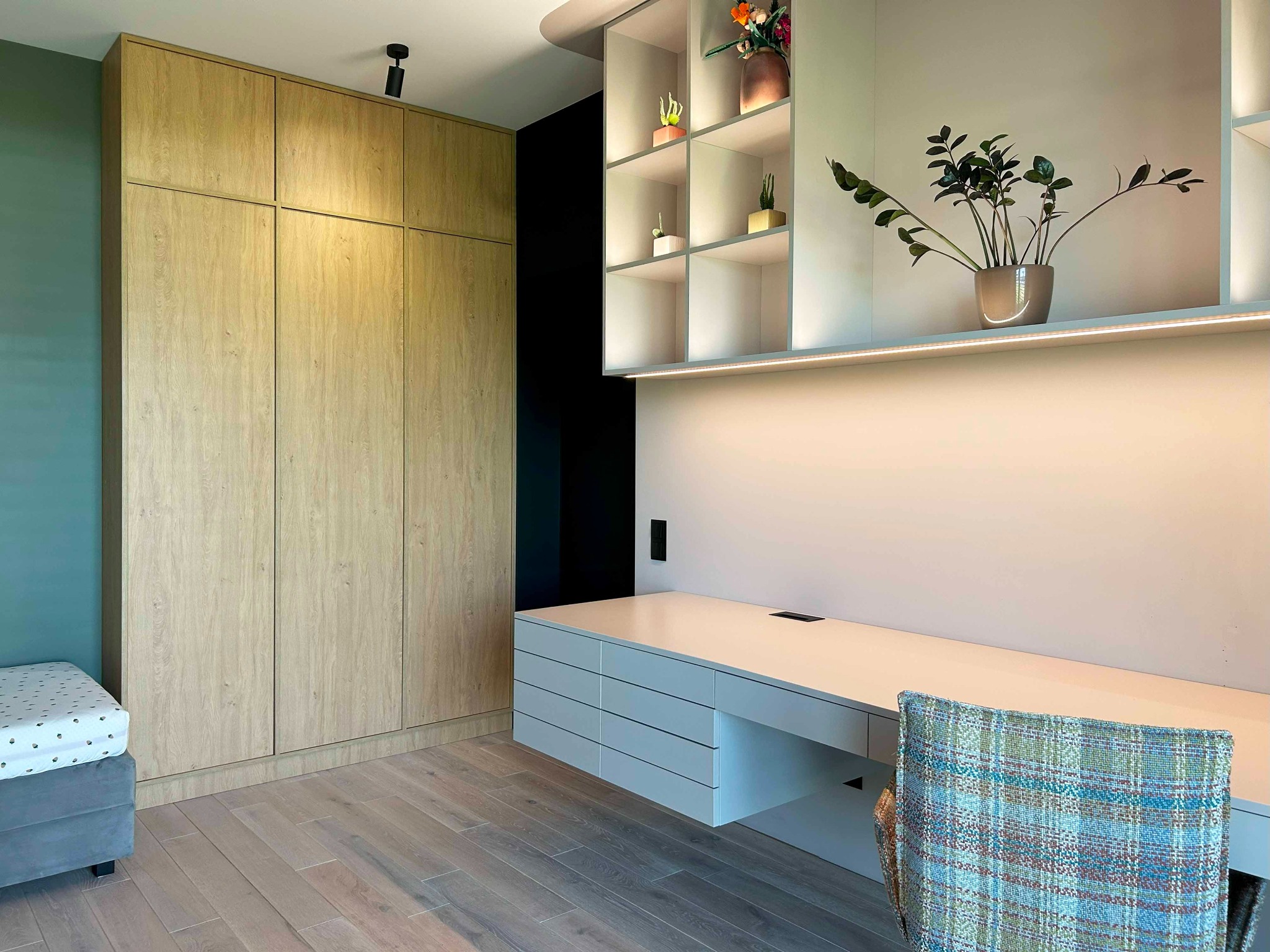 Jasna, drewniana szafa z pełnymi drzwiami obok biurka z szufladami i półkami z podświetleniem LED, na których stoją rośliny doniczkowe. W tle ciemna ściana.