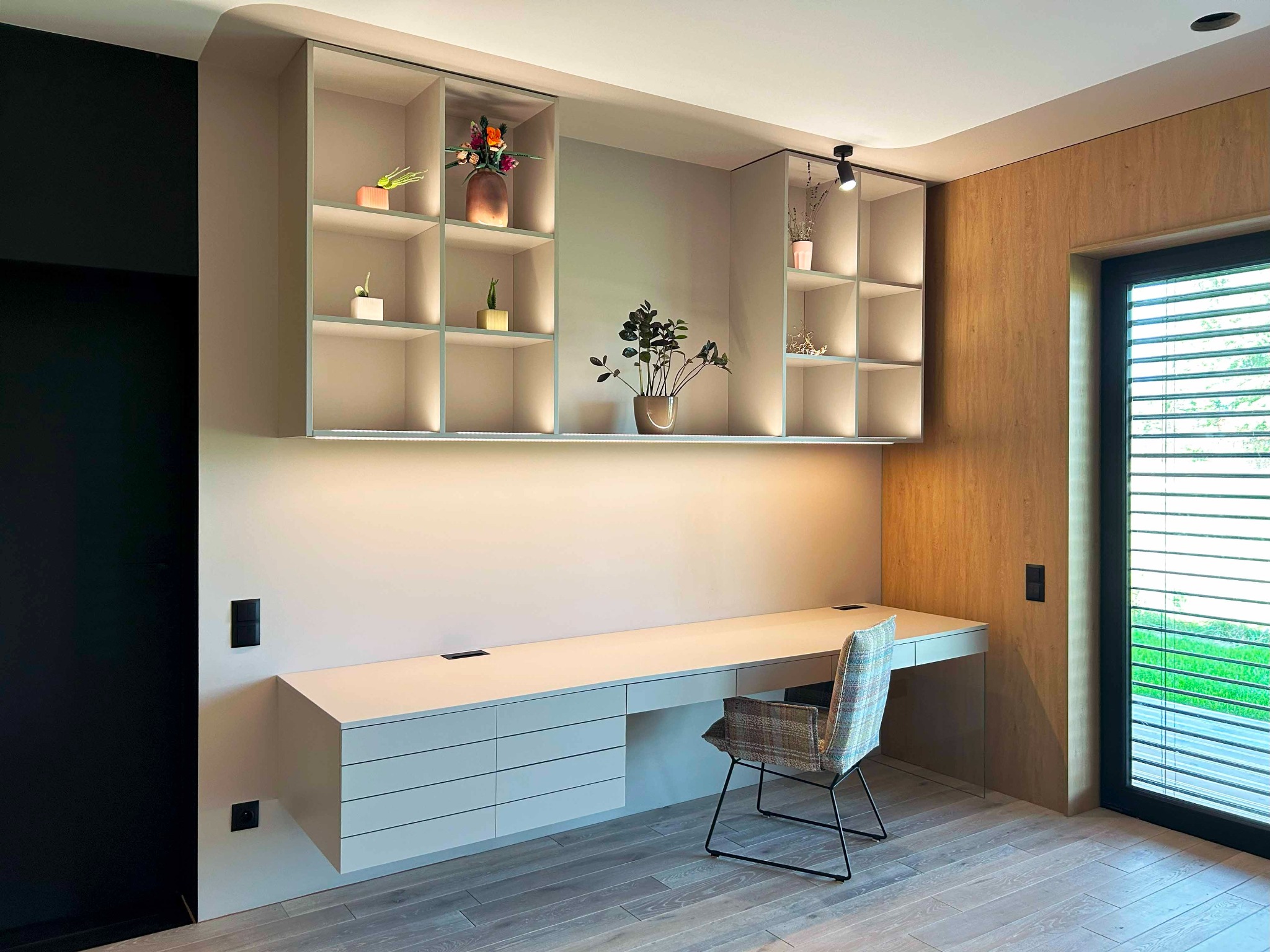 Nowoczesne biurko z szufladami i wiszącymi półkami, oświetlone LED, z dekoracjami roślinnymi, krzesło w kratę, drewniana podłoga i okno z żaluzjami.