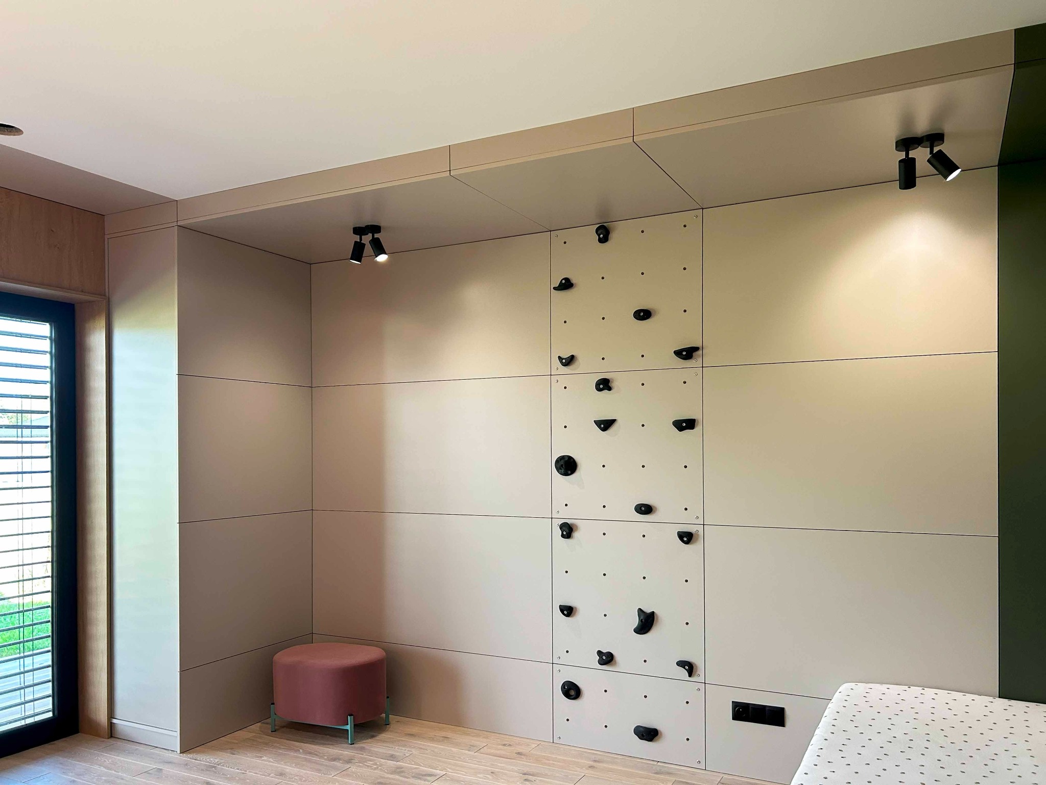 Beżowa zabudowa ściany z panelami, wbudowaną ścianką wspinaczkową z czarnymi uchwytami, oświetleniem punktowym i pufą w pokoju dziecięcym.