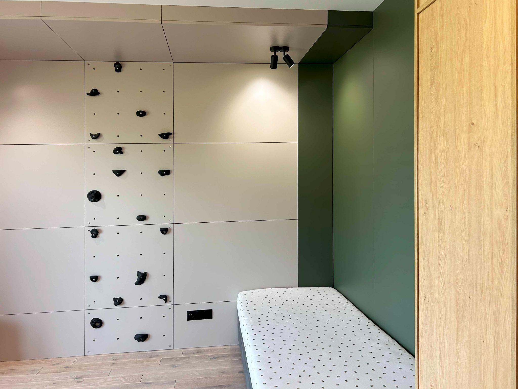Pokój dziecięcy z modułową ścianką wspinaczkową z czarnymi uchwytami, oświetleniem punktowym i tapczanem w rogu, obok drewnianej szafy.