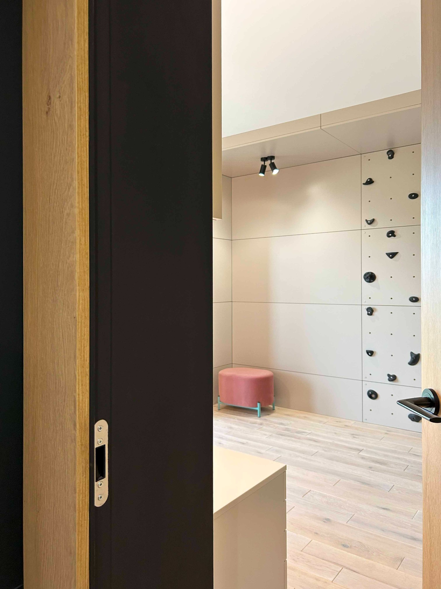 Pokój dziecięcy z ścianką wspinaczkową w neutralnych kolorach, widok przez otwarte drzwi z czarną wstawką i drewnianą ramą, różowy puf na tle