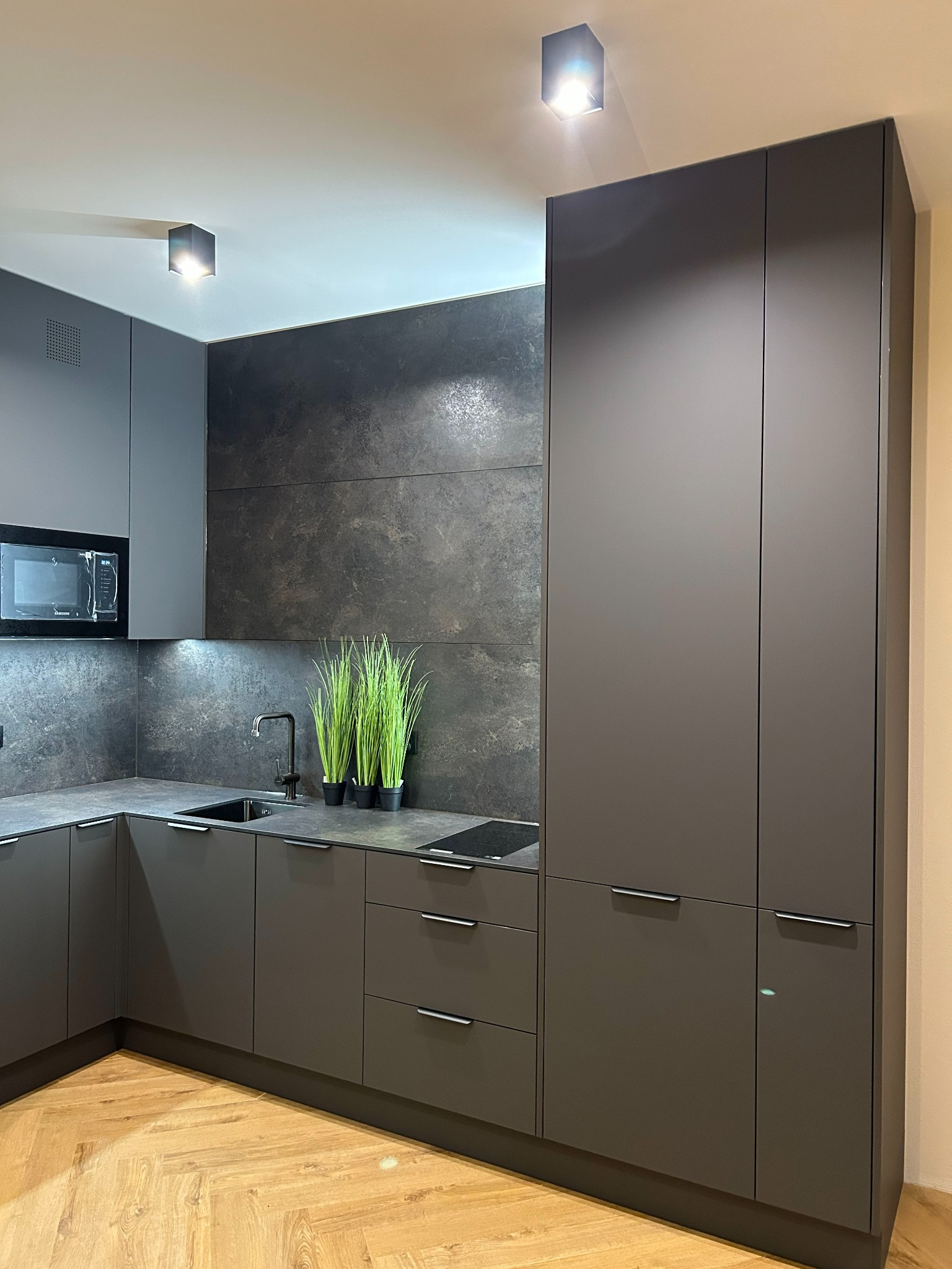 Grafitowa kuchnia z mikrofala w zabudowie, czarną armaturą i roślinami na blacie. Minimalistyczny design z ciemnym kamiennym panelem na ścianie i parkietem w jodełkę.
