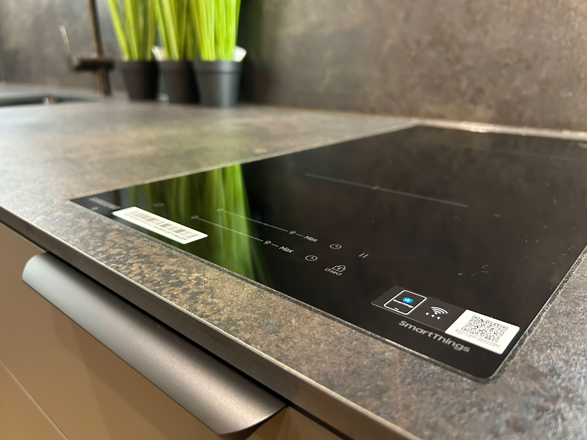 Indukcyjna płyta grzewcza SmartThings wkomponowana w blat kuchenny o szarej, nakrapianej fakturze. W tle doniczki z zielonymi roślinami.