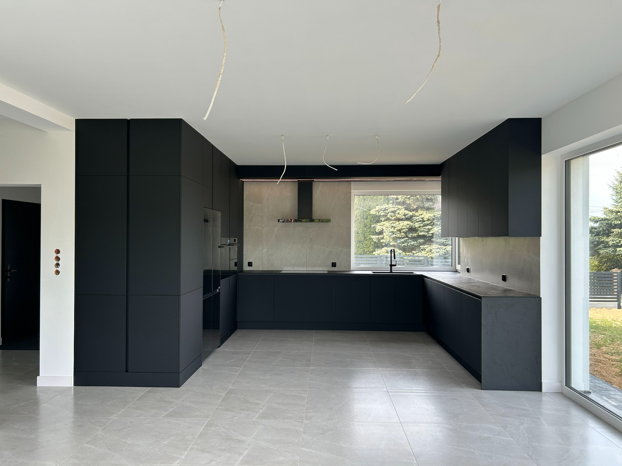 Nowoczesna, czarna kuchnia w kształcie litery U z szarymi płytkami i dużym oknem z widokiem na zieleń. Minimalistyczny design z wiszącymi kablami.
