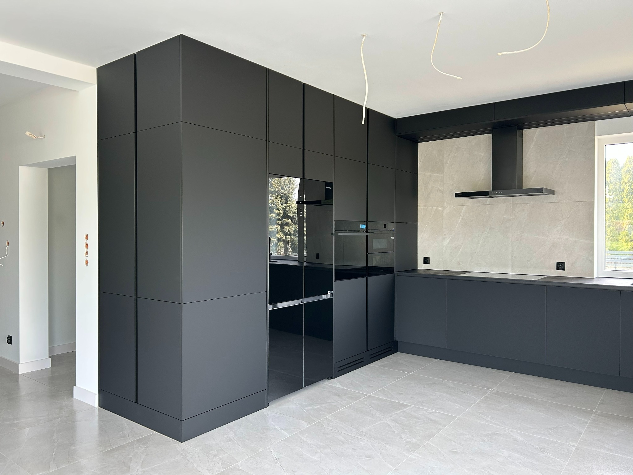 Nowoczesna, czarna kuchnia z zabudową po sufit i szklanym frontem lodówki, na tle marmurowej ściany i jasnej podłogi. Widoczne niedokończone kable oświetleniowe.