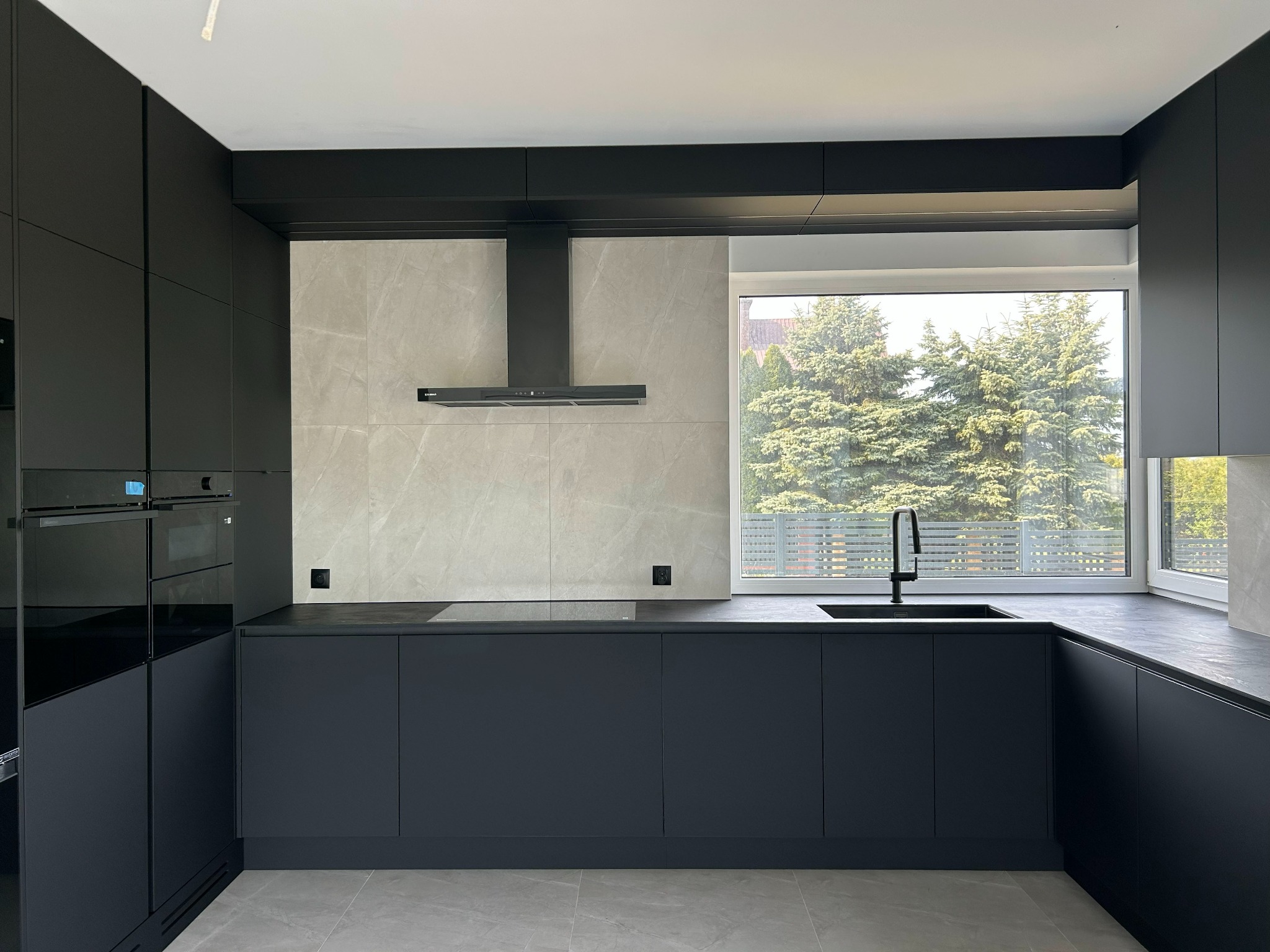 Nowoczesna, minimalistyczna kuchnia w odcieniach szarości i czerni. Matowe fronty szafek, marmurowe płytki na ścianie, okno z widokiem na zieleń. Elegancki design.