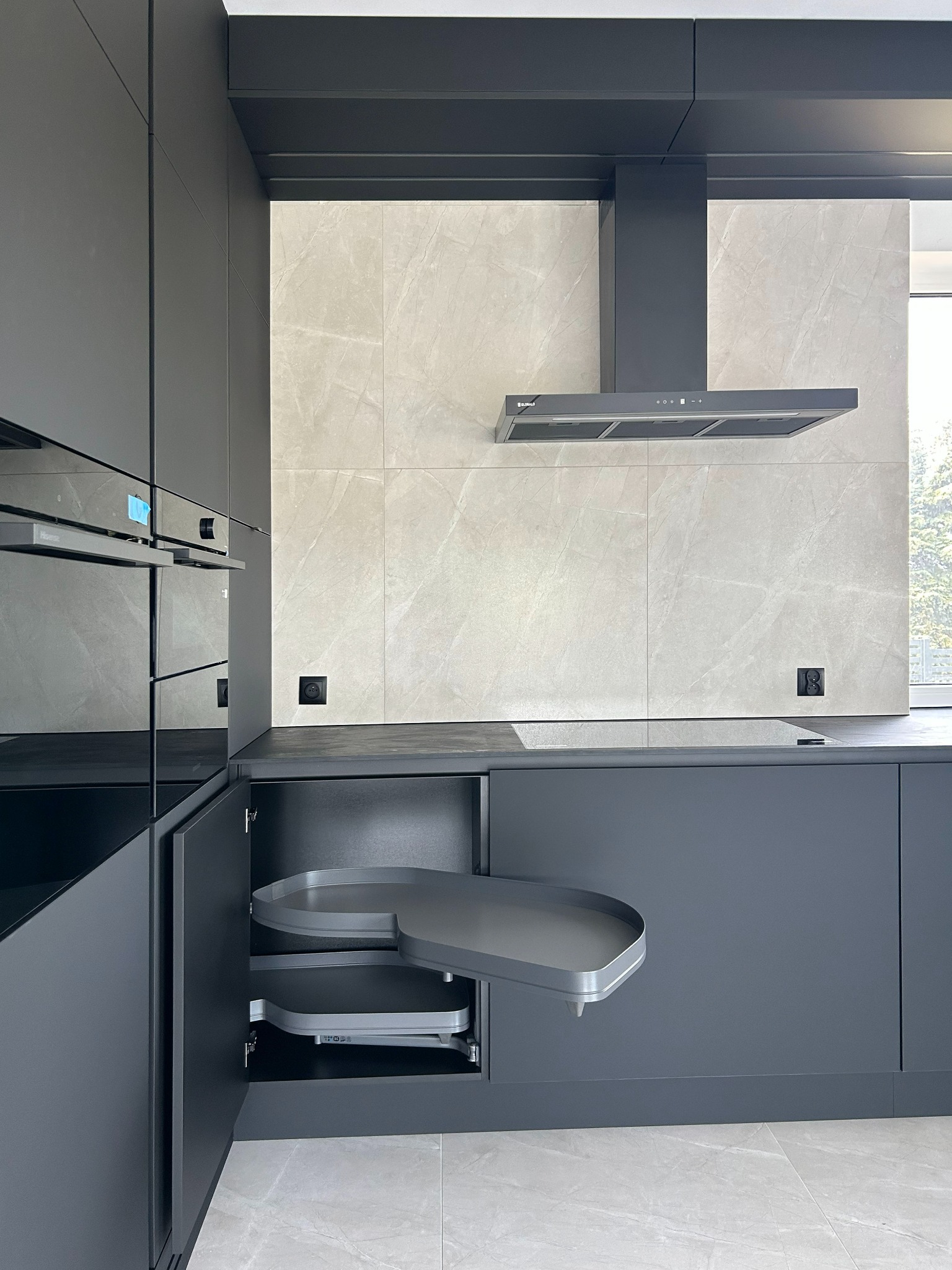 Nowoczesna, grafitowa kuchnia z otwartą szafką narożną typu 'nerka', płyta indukcyjna i okap. Minimalistyczny design, marmurowe płytki na ścianie.