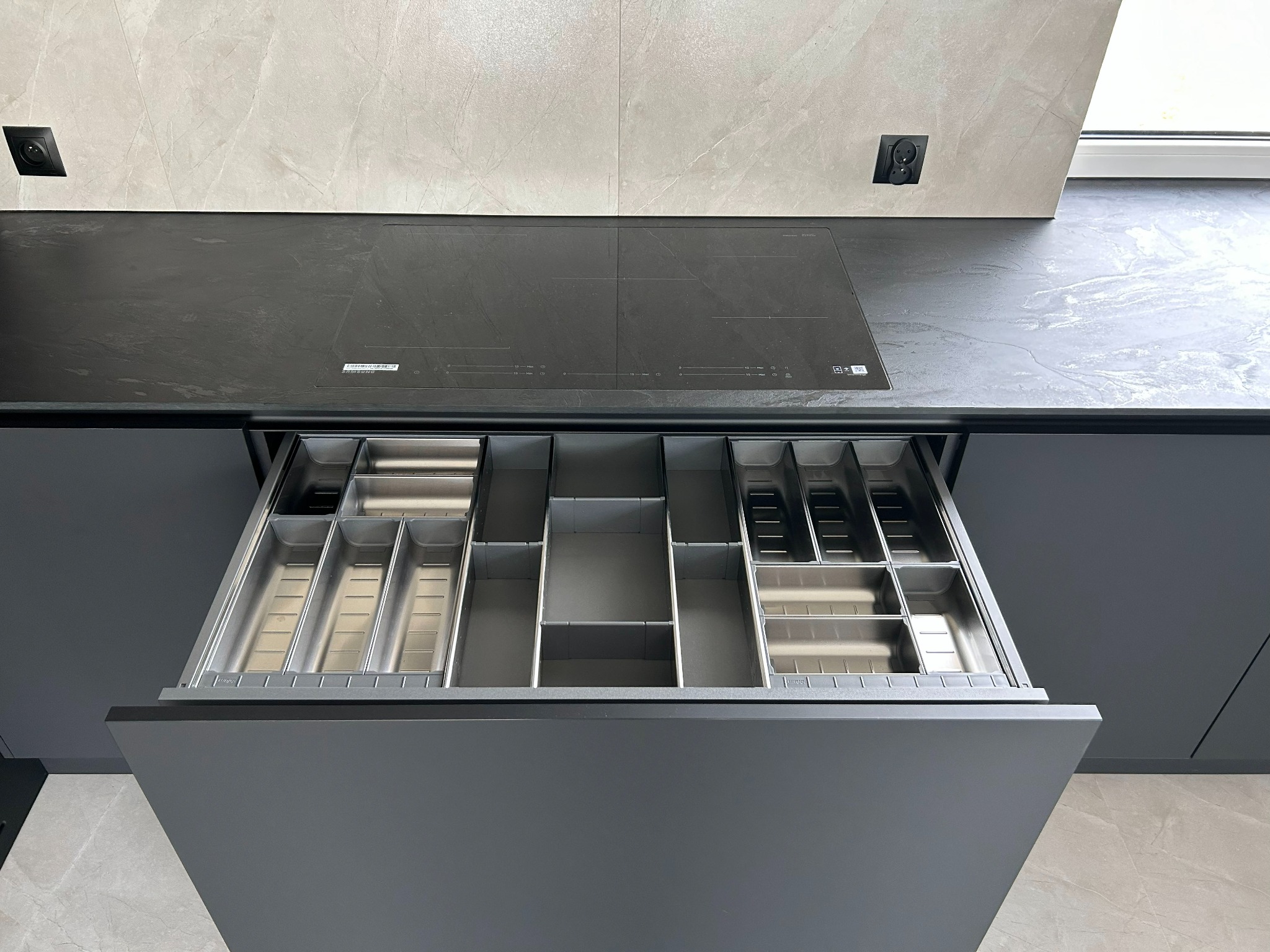 Otwarta szuflada z metalowymi organizerami na sztućce w nowoczesnej, szarej kuchni z czarnym blatem i płytą indukcyjną. Minimalistyczny design, jasne płytki na ścianie.