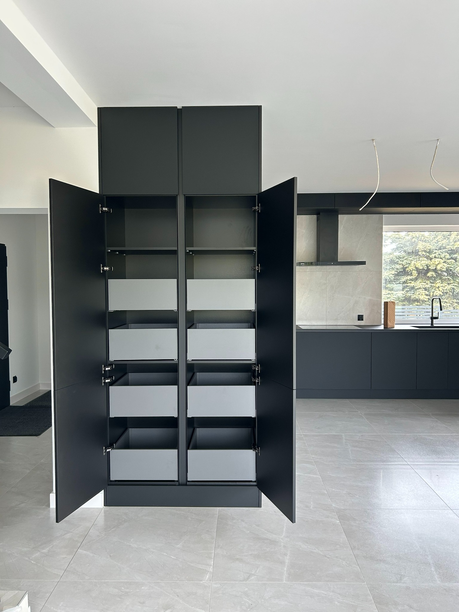 Nowoczesna, czarna szafa kuchenna z otwartymi drzwiami i szufladami w odcieniach szarości, na tle jasnej kuchni z oknem i widokiem na zieleń.