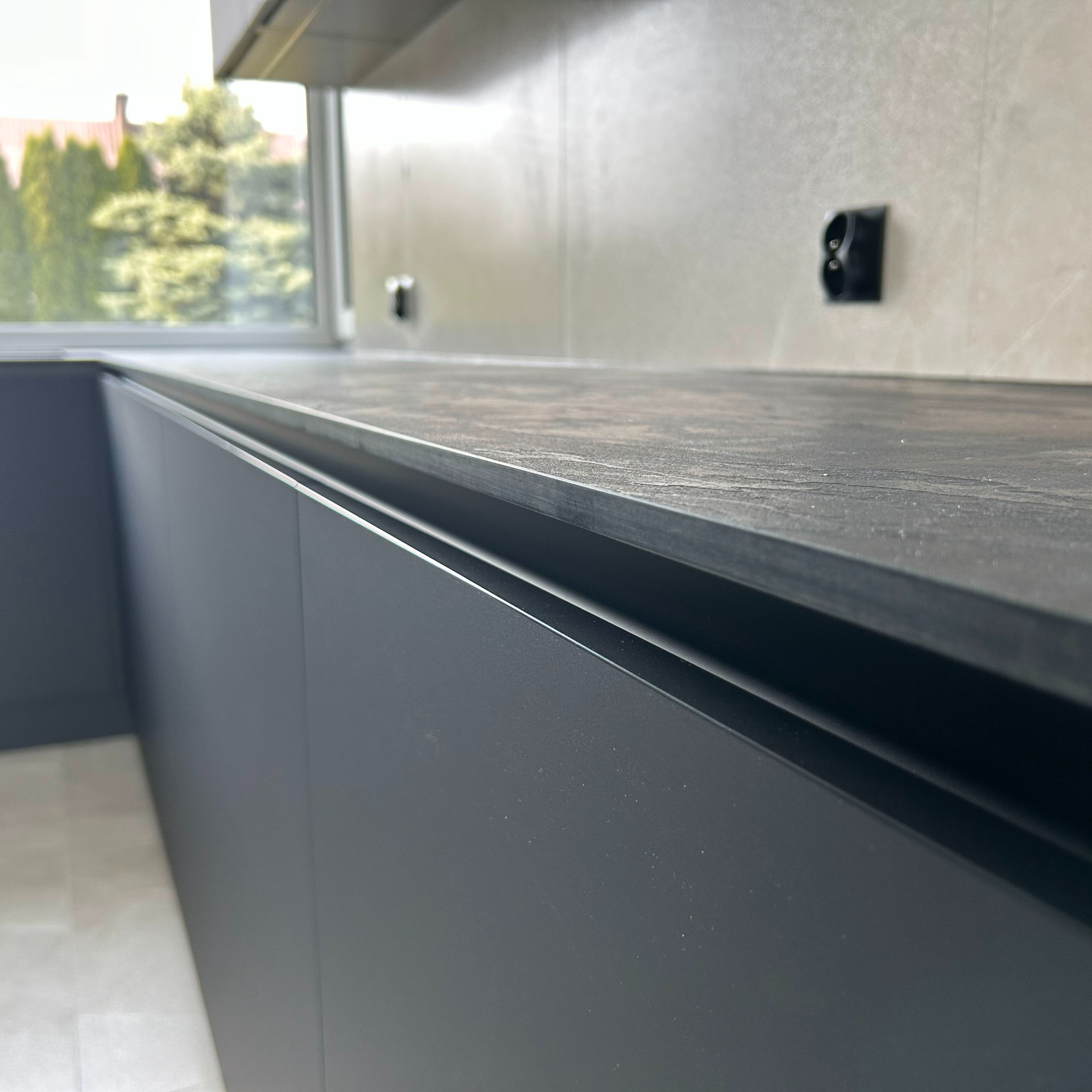 Szary blat kuchenny z widocznym gniazdkiem elektrycznym na ścianie i oknem w tle, prezentujący nowoczesny design i dbałość o detale wykończenia.