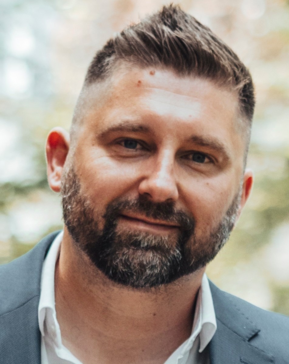 Portret mężczyzny z brodą, w szarej marynarce i białej koszuli, uśmiechającego się do kamery na rozmytym tle.