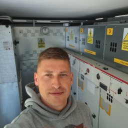 Mariusz Góral - Mężczyzna w szarej bluzie z kapturem robiący selfie na tle rozdzielnicy elektrycznej z żółtymi tabliczkami ostrzegawczymi.