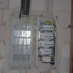 Mariusz Góral - Dwa rozdzielnice elektryczne w trakcie instalacji w Skierniewicach, z widocznymi przewodami, zamontowane na surowej ścianie z betonu komórkowego.