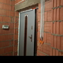 Surowe ściany z czerwonej cegły z widocznymi przewodami elektrycznymi w białej osłonie, prowadzącymi do pomarańczowych puszek instalacyjnych, nad szarą futryną drzwiową w trakcie montażu.