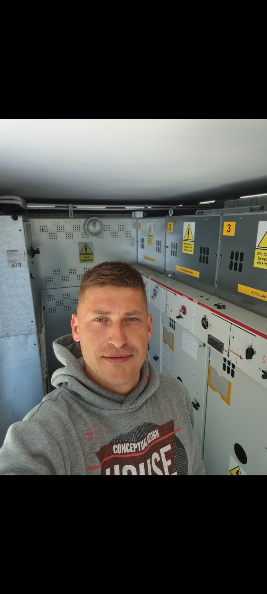Mężczyzna w szarej bluzie z kapturem robiący selfie na tle rozdzielnicy elektrycznej z żółtymi tabliczkami ostrzegawczymi.