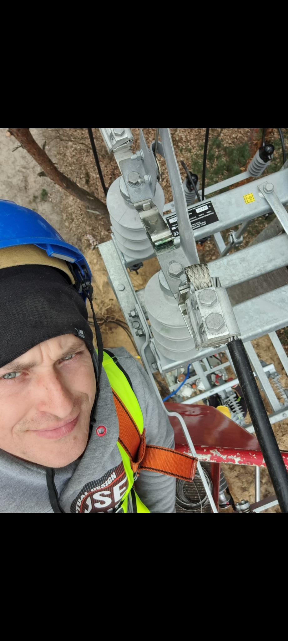 Elektryk w kasku i uprzęży, z bliska, na tle konstrukcji wsporczej z izolatorami i przewodami wysokiego napięcia.