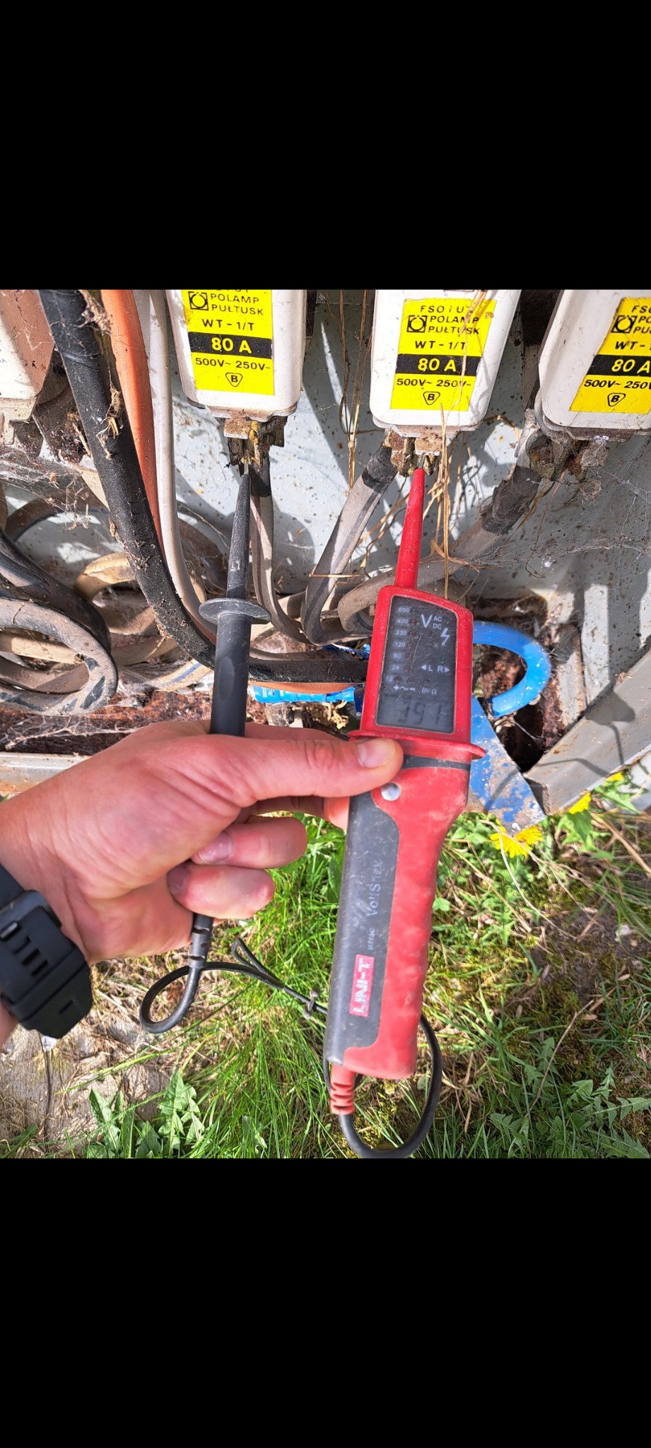 Pomiar napięcia na zabezpieczeniach instalacji elektrycznej przy użyciu miernika Unit-T VoltStick, widoczne bezpieczniki POLAMP Pułtusk 80A.