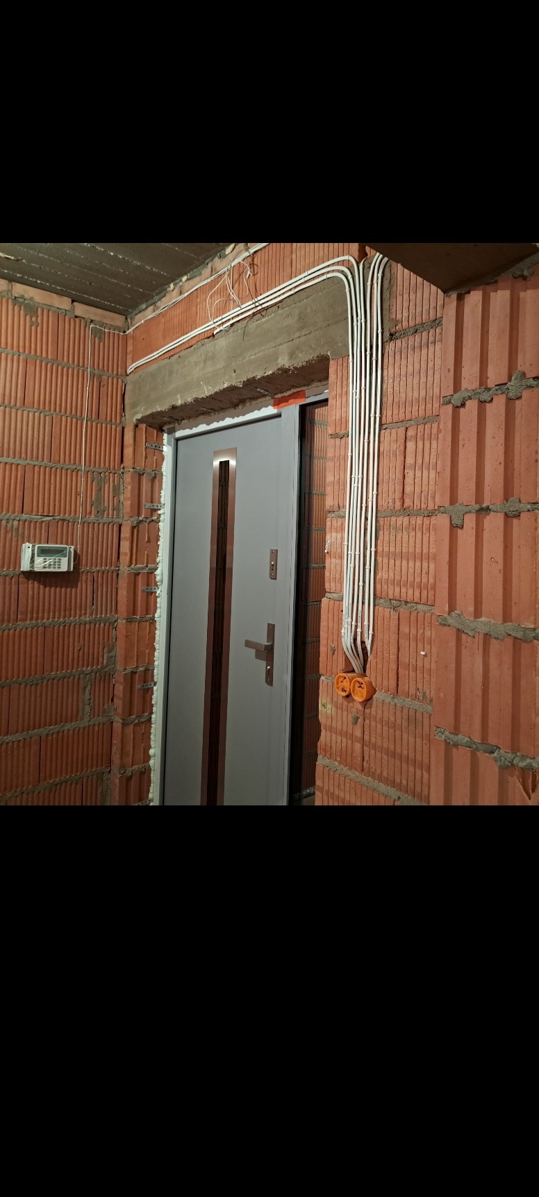 Surowe ściany z czerwonej cegły z widocznymi przewodami elektrycznymi w białej osłonie, prowadzącymi do pomarańczowych puszek instalacyjnych, nad szarą futryną drzwiową w trakcie montażu.