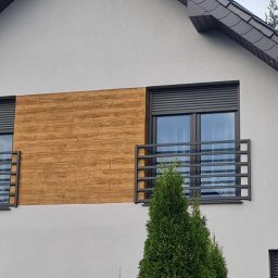 GAPEX Kamil Gapiński - Szara elewacja domu z dwoma oknami balkonowymi zabezpieczonymi metalowymi balustradami w kolorze grafitowym, z roletami zewnętrznymi i okładziną drewnopodobną pomiędzy oknami.