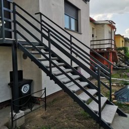 GAPEX Kamil Gapiński - Zewnętrzne schody z czarnej stali z szarymi stopniami prowadzące do wejścia do budynku, obok śmietnika z graffiti.