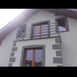 GAPEX Kamil Gapiński - Szary dom jednorodzinny z balkonem zabezpieczonym prostą, poziomą, metalową konstrukcją, widoczne okna z brązowymi ramami i dekoracyjne szare kwadraty na elewacji.