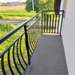 Czarna, metalowa balustrada balkonowa z zakrzywionymi elementami w Budzyniu, widok na pola i zabudowania w tle.
