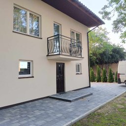 GAPEX Kamil Gapiński - Dwukondygnacyjny dom z beżową elewacją, czarną balustradą balkonową z dekoracyjnymi prętami, ciemnymi oknami i drzwiami wejściowymi, wyłożony szarą kostką brukową, z widocznym fragmentem białego...