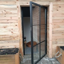 GAPEX Kamil Gapiński - Otwarta, przeszklona, czarna metalowa rama drzwiowa w drewnianej ścianie budynku, widoczne donice z ziemią i sznurkowymi uchwytami po bokach.