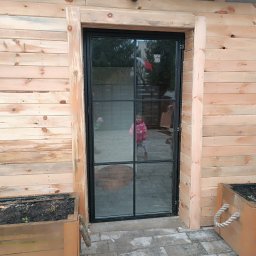 GAPEX Kamil Gapiński - Nowoczesne drzwi z czarną metalową ramą i szklanymi panelami, wkomponowane w drewnianą elewację budynku z widocznymi skrzyniami na rośliny po bokach.