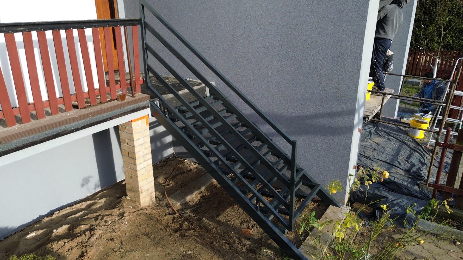 Zewnętrzne metalowe schody prowadzące do budynku, świeżo pomalowane na grafitowo, z widocznymi śladami prac malarskich w tle i kubkiem na balustradzie balkonu.