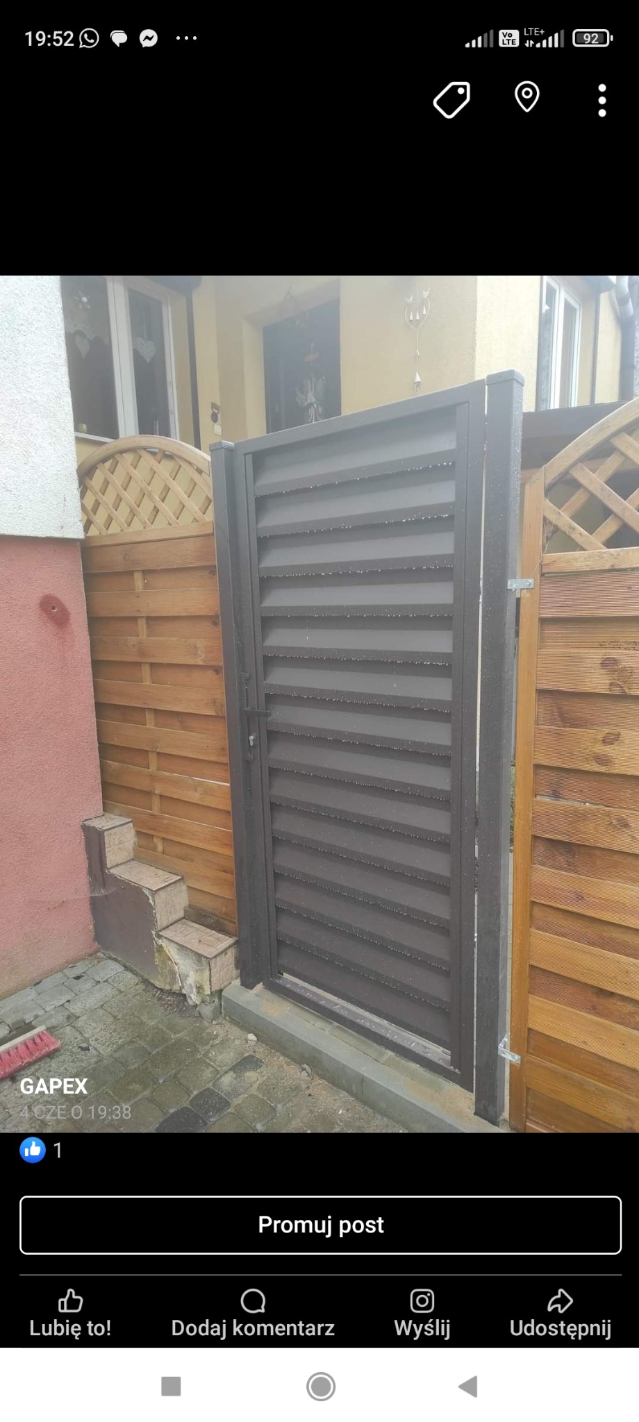 Nowoczesna, brązowa furtka z poziomymi lamelami, zamontowana przy drewnianym ogrodzeniu z dekoracyjną kratką, widoczna z bliska na tle jasnego budynku.