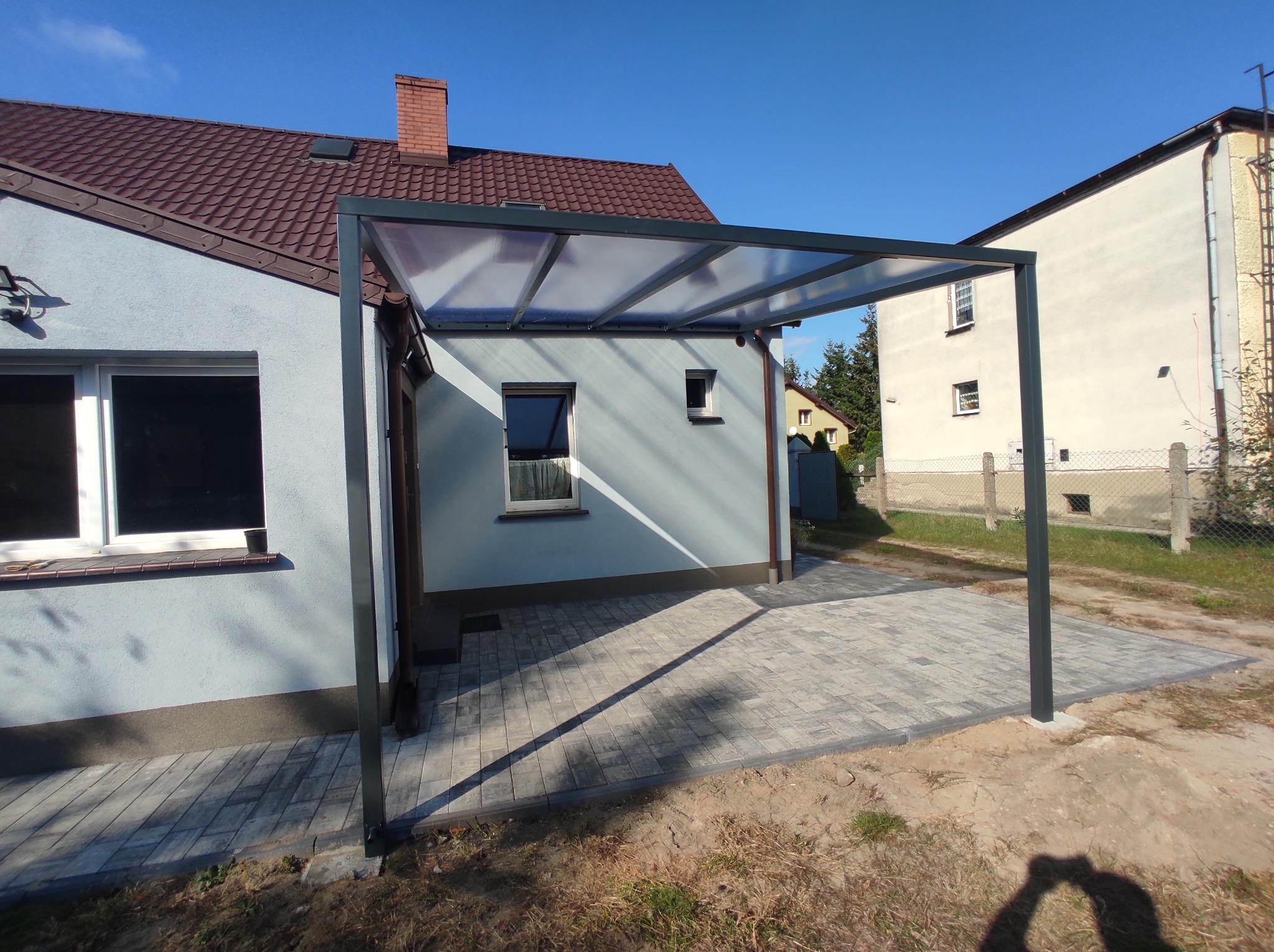 Nowoczesna, szara pergola z przezroczystym zadaszeniem, przylegająca do jasnoniebieskiego domu z brązowym dachem, z kostką brukową pod spodem i widocznym fragmentem drugiego budynku w tle.