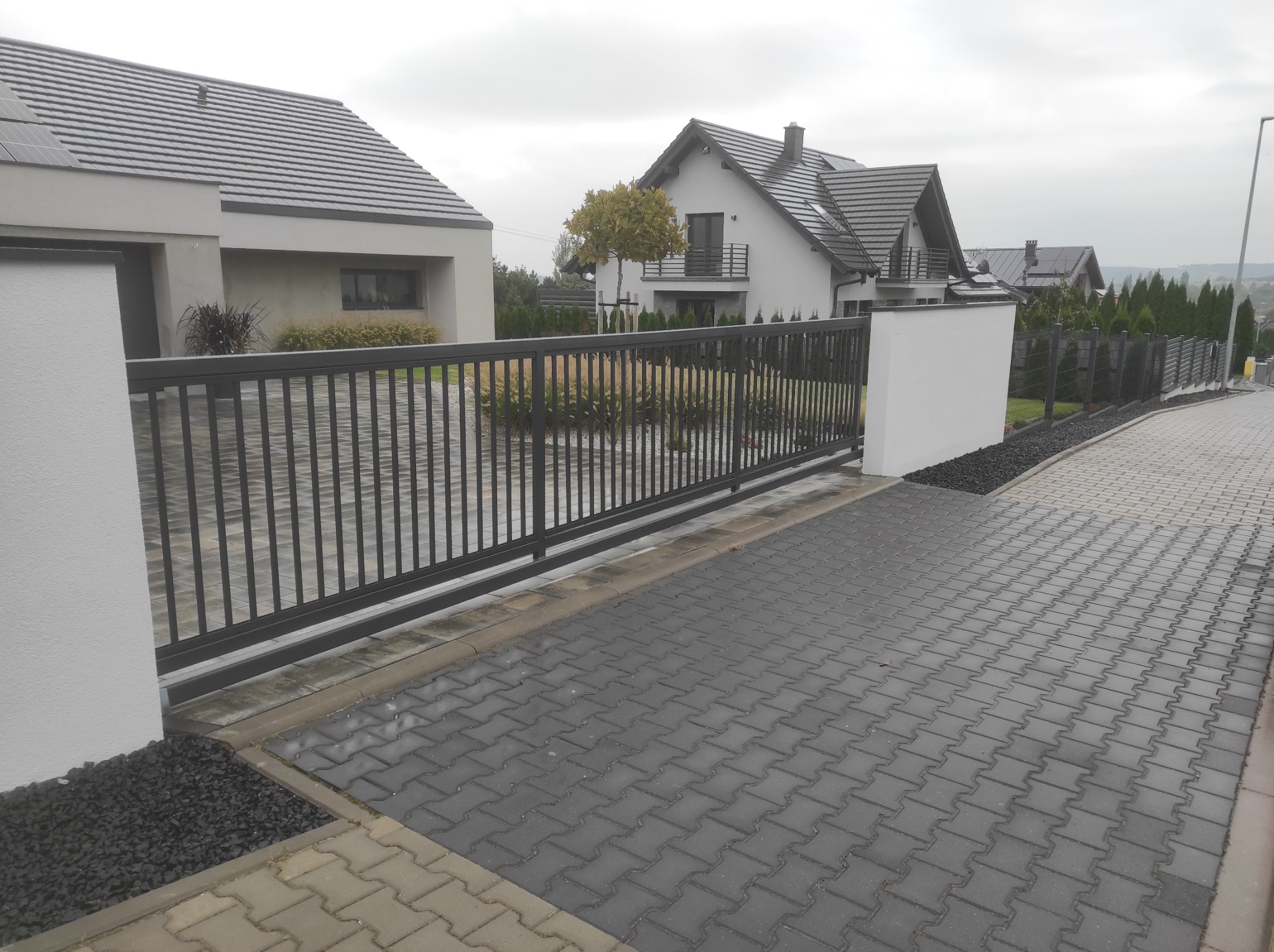 Nowoczesna posesja z przesuwną bramą i ogrodzeniem z metalowych sztachet w kolorze grafitowym, wykończona białymi słupkami, bruk w szarym odcieniu, w tle domy jednorodzinne z szarymi dachami.