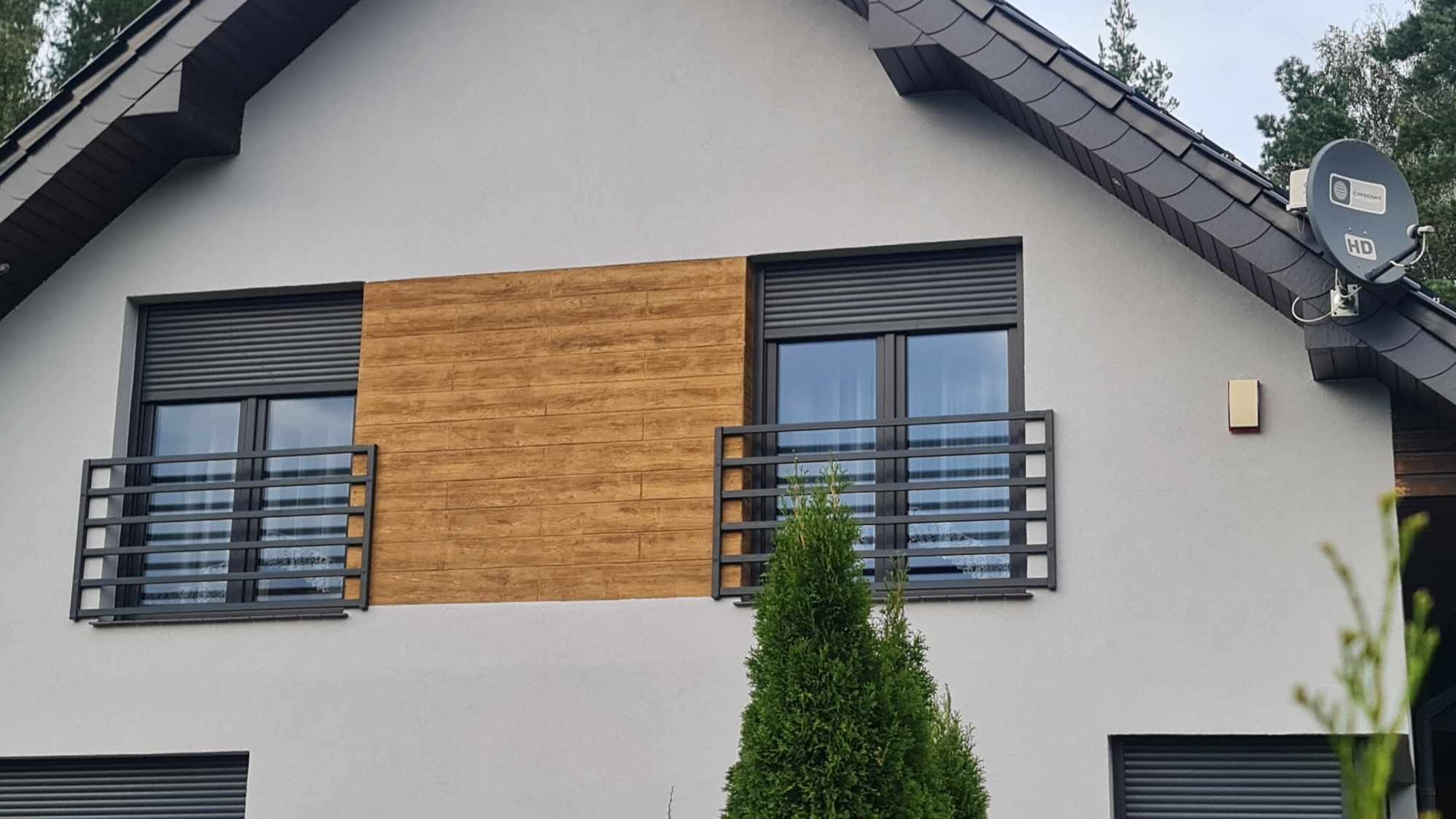 Szara elewacja domu z dwoma oknami balkonowymi zabezpieczonymi metalowymi balustradami w kolorze grafitowym, z roletami zewnętrznymi i okładziną drewnopodobną pomiędzy oknami.