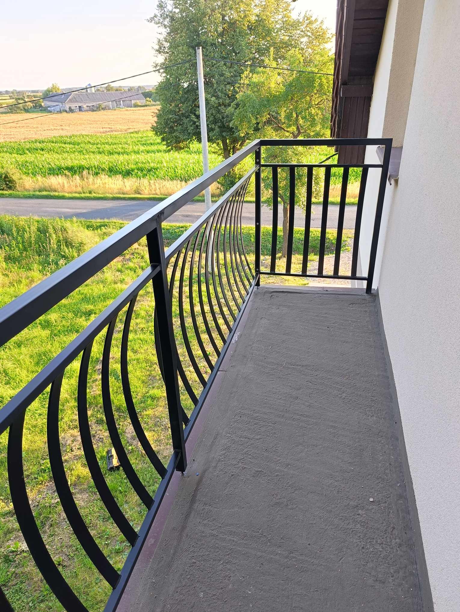 Czarna, metalowa balustrada balkonowa z zakrzywionymi elementami w Budzyniu, widok na pola i zabudowania w tle.