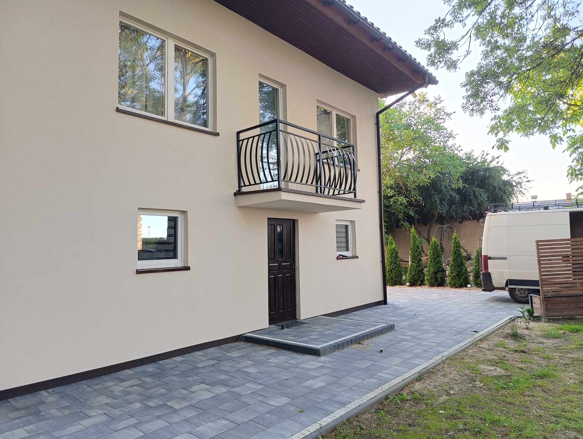 Dwukondygnacyjny dom z beżową elewacją, czarną balustradą balkonową z dekoracyjnymi prętami, ciemnymi oknami i drzwiami wejściowymi, wyłożony szarą kostką brukową, z widocznym fragmentem białego...